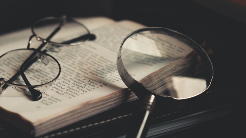 An open book with glasses and a magnifying glass resting on its pages, symbolizing close reading of investigative books