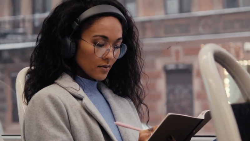 Woman with headphones listening to an audiobook and writing in a notebook during her commute