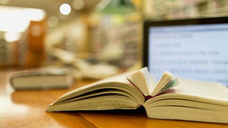 Open book on a desk in front of a laptop screen in a library, symbolizing how to track your reading