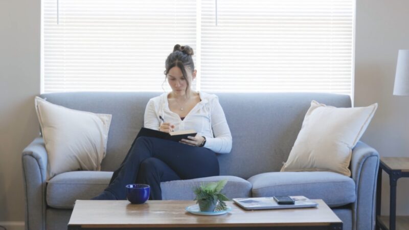 Woman writes notes in a journal on a sofa in a sunlit room